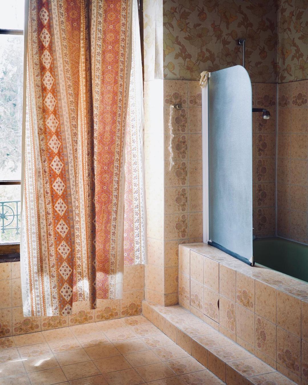 Bathroom interior featuring patterned tile walls and floors, a curtained window, and a bathtub with a glass screen in natural light.
