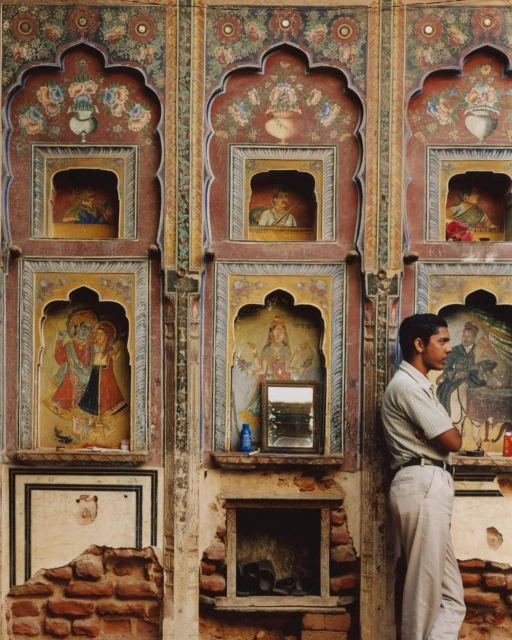 Adult man stands in front of a colorful brick wall with archways and painted artwork in an outdoor setting during daytime.