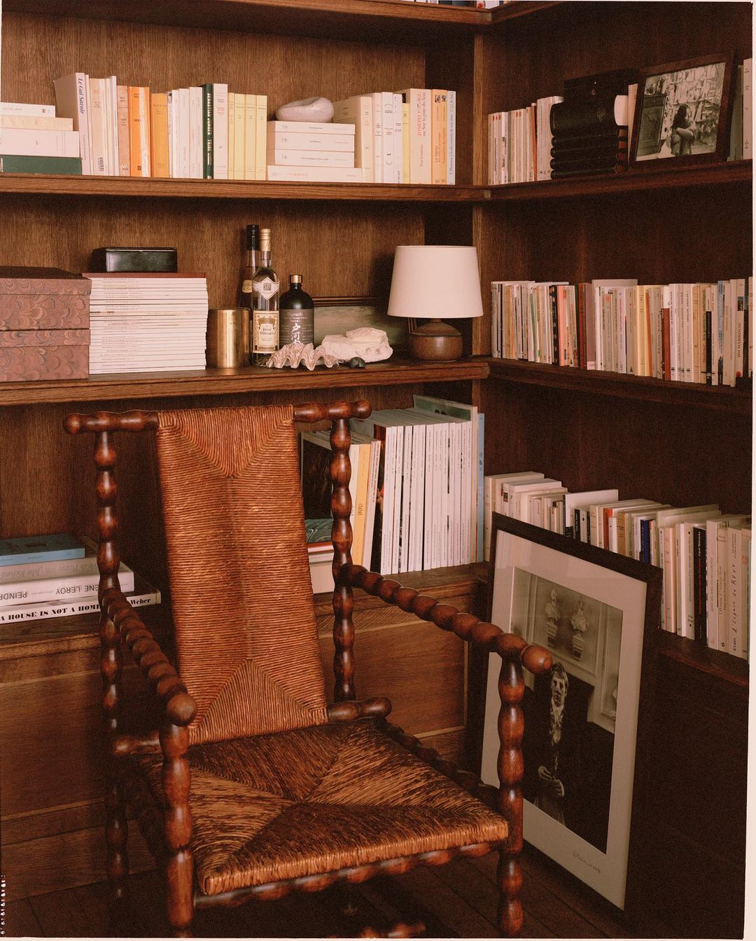 Interior view of a home library with a woven armchair in the foreground, bookshelves filled with books, and decorative objects.