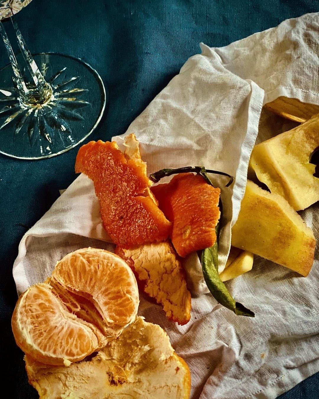 Still life featuring a peeled tangerine, apple peels, and a crystal wine glass atop a white cloth against a dark blue background.