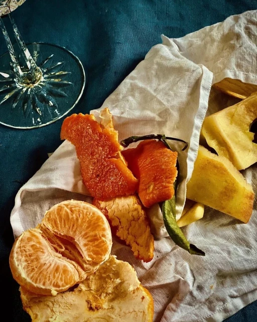 Still life featuring a peeled tangerine, apple peels, and a crystal wine glass atop a white cloth against a dark blue background.