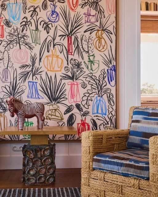 Living room interior with a straw armchair, wood side table holding a zebra figure, patterned rug, and colorful plant print wallpaper.