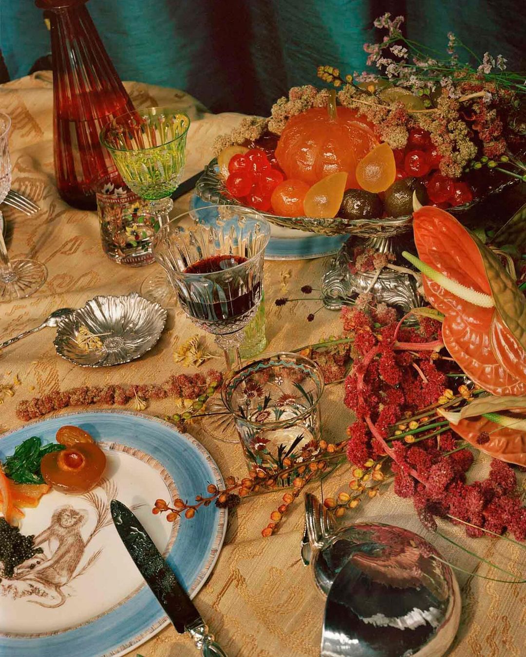 Artful still life on a table with desserts, wine glasses, ornate flatware, antique dinnerware, flowers and a red bottle.