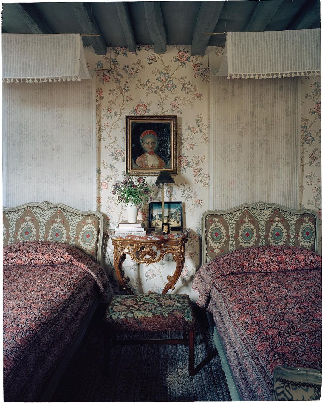 Bedroom interior with two ornate beds, floral wallpaper, marble-topped table, and framed portrait in a traditional style.