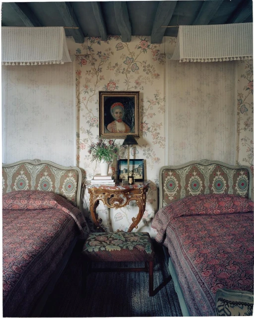Bedroom interior with two ornate beds, floral wallpaper, marble-topped table, and framed portrait in a traditional style.
