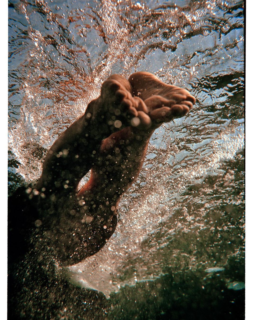 Underwater view of adult human feet and legs submerged in water, creating splash effect in clear liquid with visible sun rays.
