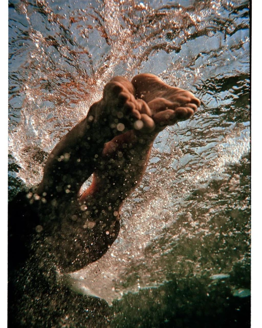 Underwater view of adult human feet and legs submerged in water, creating splash effect in clear liquid with visible sun rays.