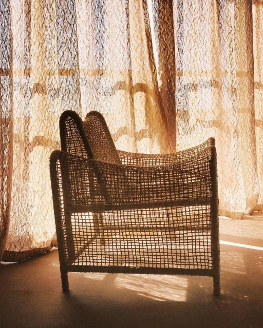 Woven wicker loveseat armchair is positioned in front of semi-sheer patterned curtains with natural light streaming through the window.