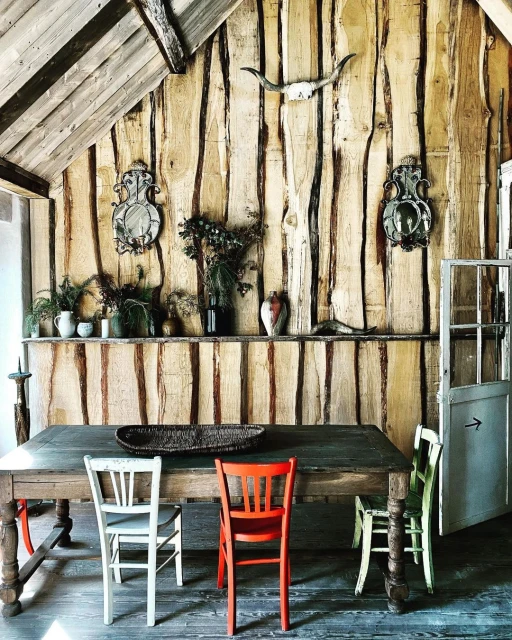 Rustic dining room featuring a wooden table with colorful chairs, set against a backdrop of textured wooden walls, plants, and natural light.