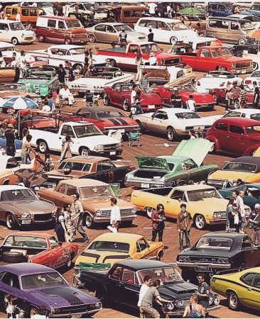 High-angle shot of a crowded outdoor car meet, with various classic automobiles parked and people gathered around them in daylight.