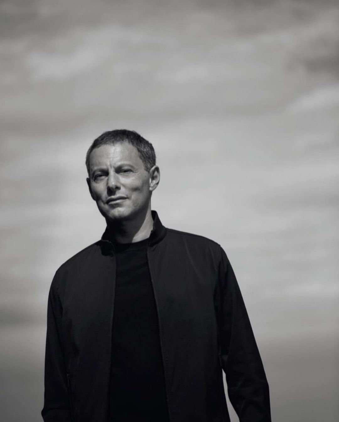 Black and white portrait of a middle-aged adult male in a black jacket against a cloudy sky, captured in a close-up shot.