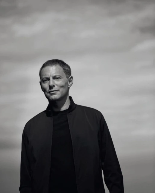 Black and white portrait of a middle-aged adult male in a black jacket against a cloudy sky, captured in a close-up shot.