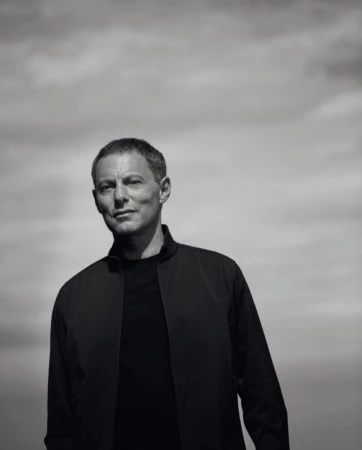 Black and white portrait of a middle-aged adult male in a black jacket against a cloudy sky, captured in a close-up shot.