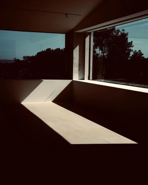 Sunlight streams into an empty, modern room through large windows, highlighting the neutral-toned floor and architectural lines.