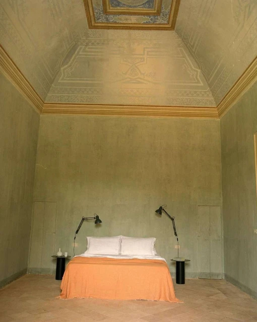 Interior of a bedroom features a bed with an orange blanket, flanked by tables and lamps, against a pale green wall with ceiling design.
