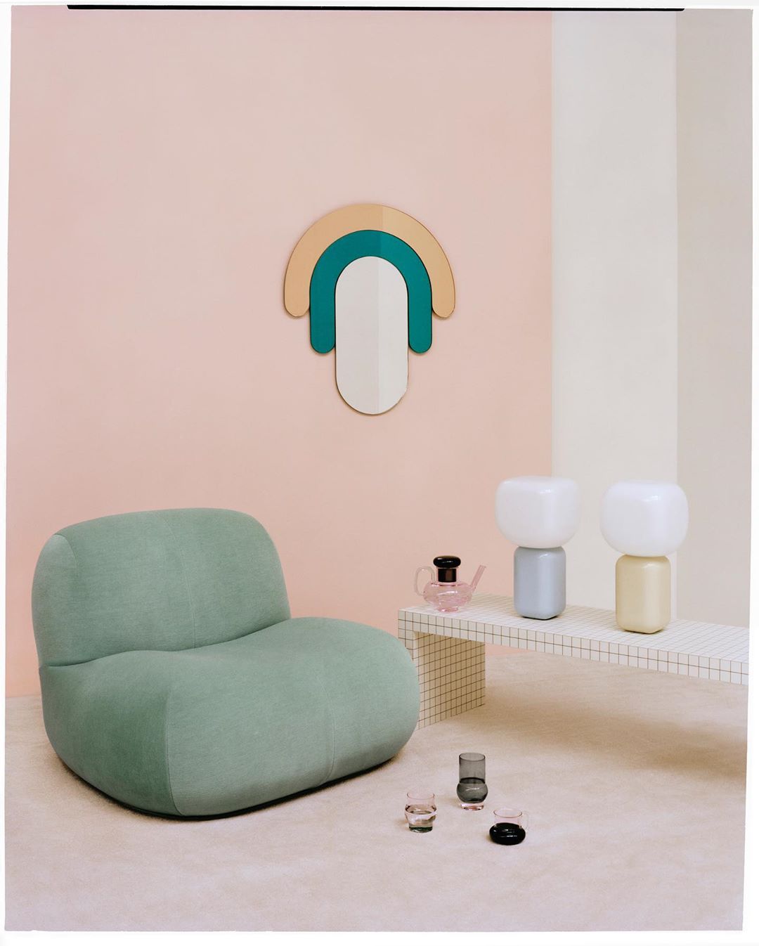 Interior design scene featuring a green armchair, side table with decor lamps, a pink teapot, and a geometric wall decoration.