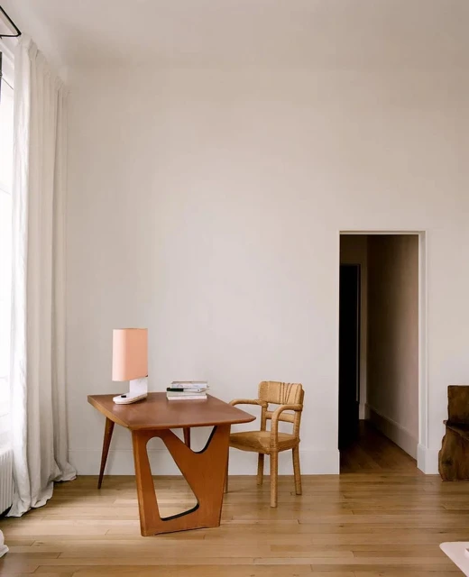 Minimalist interior with wooden desk, lamp, books, woven chair, and hardwood floor in a bright, light-filled room with neutral walls.