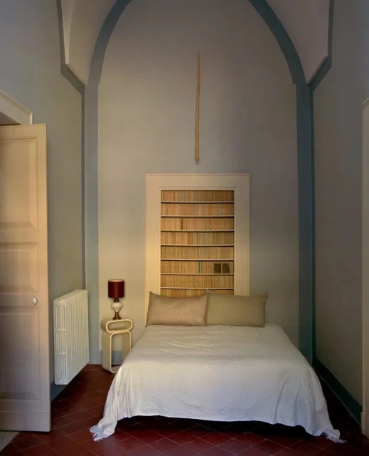 Bedroom features a bed with white linens, bookshelves as a headboard, pale blue walls, and terracotta tile flooring in a minimalist style.