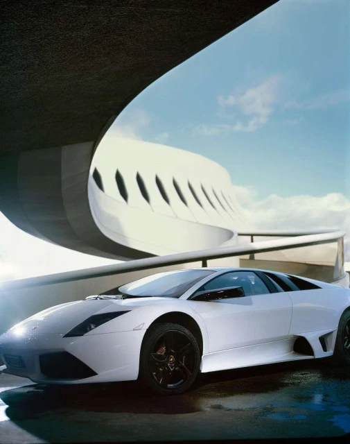 White Lamborghini Murciélago parked in an urban setting with contemporary architecture and a partially cloudy sky in the background.