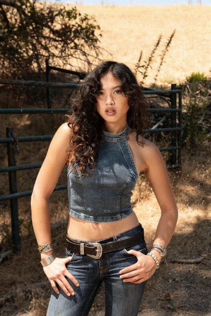 Young woman in denim crop top and jeans poses in an outdoor setting with a metal gate and grassy hillside in the background.