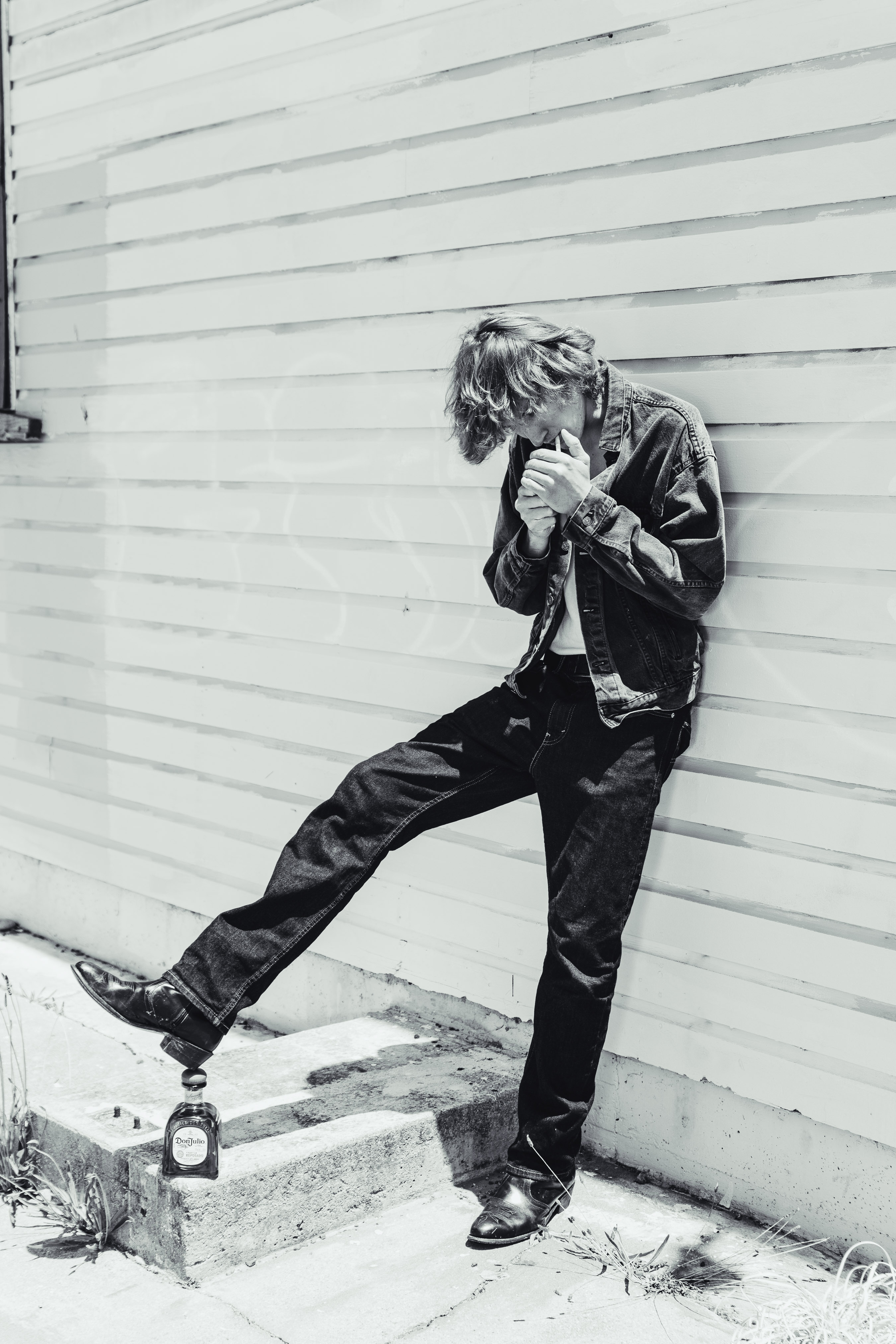 Black and white portrait of a young adult lighting a cigarette, leaning against a wall with a bottle on the ground.
