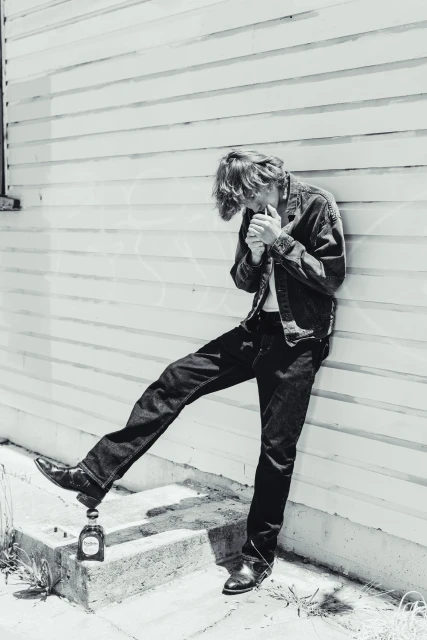 Black and white portrait of a young adult lighting a cigarette, leaning against a wall with a bottle on the ground.