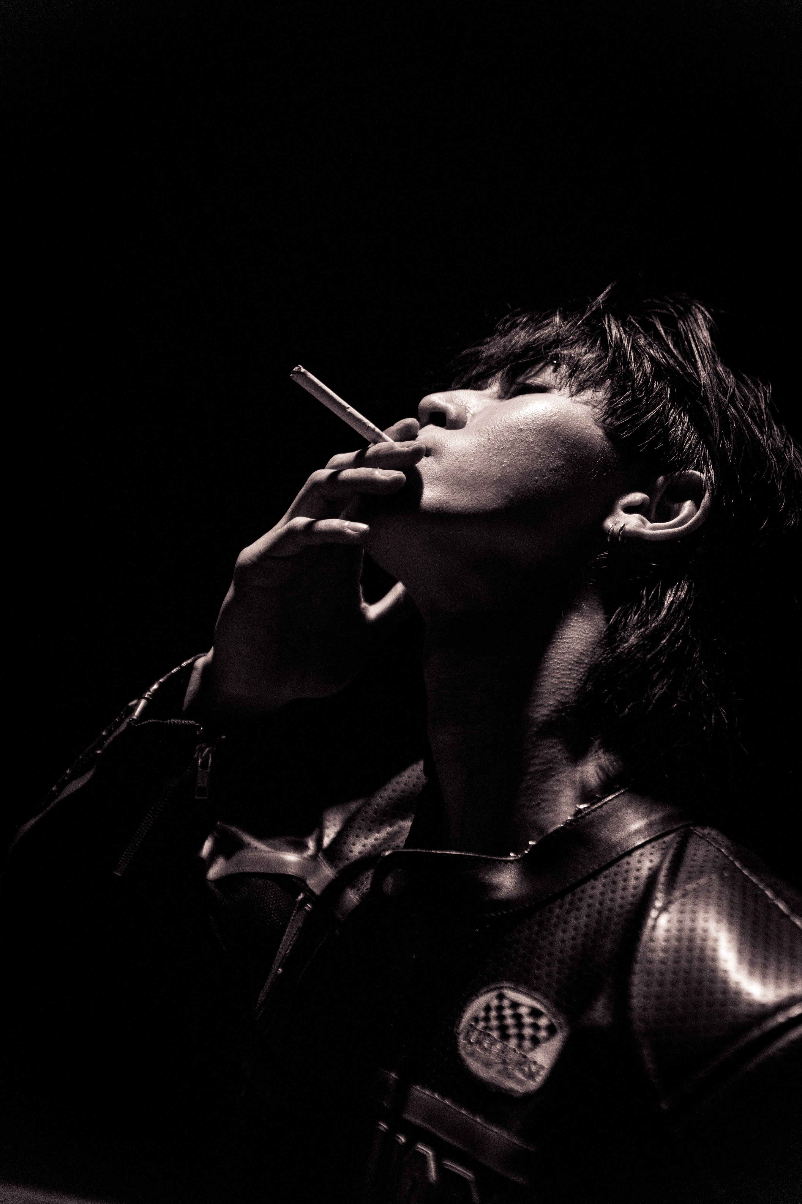 Monochrome portrait of a young adult with dark hair holding a lit cigarette looking upwards against a dark background with visible lighting.
