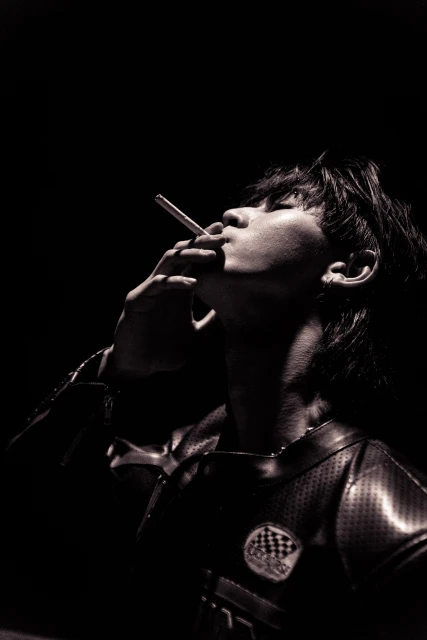Monochrome portrait of a young adult with dark hair holding a lit cigarette looking upwards against a dark background with visible lighting.