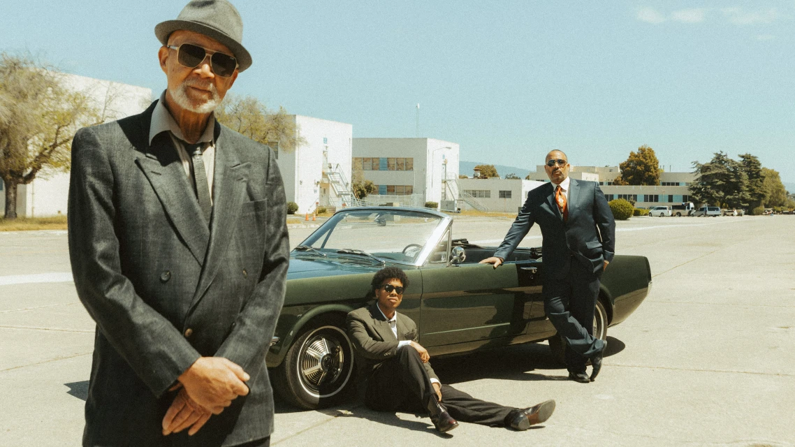 Three men in suits pose with a classic convertible car outdoors in bright daylight with a building visible in the background.