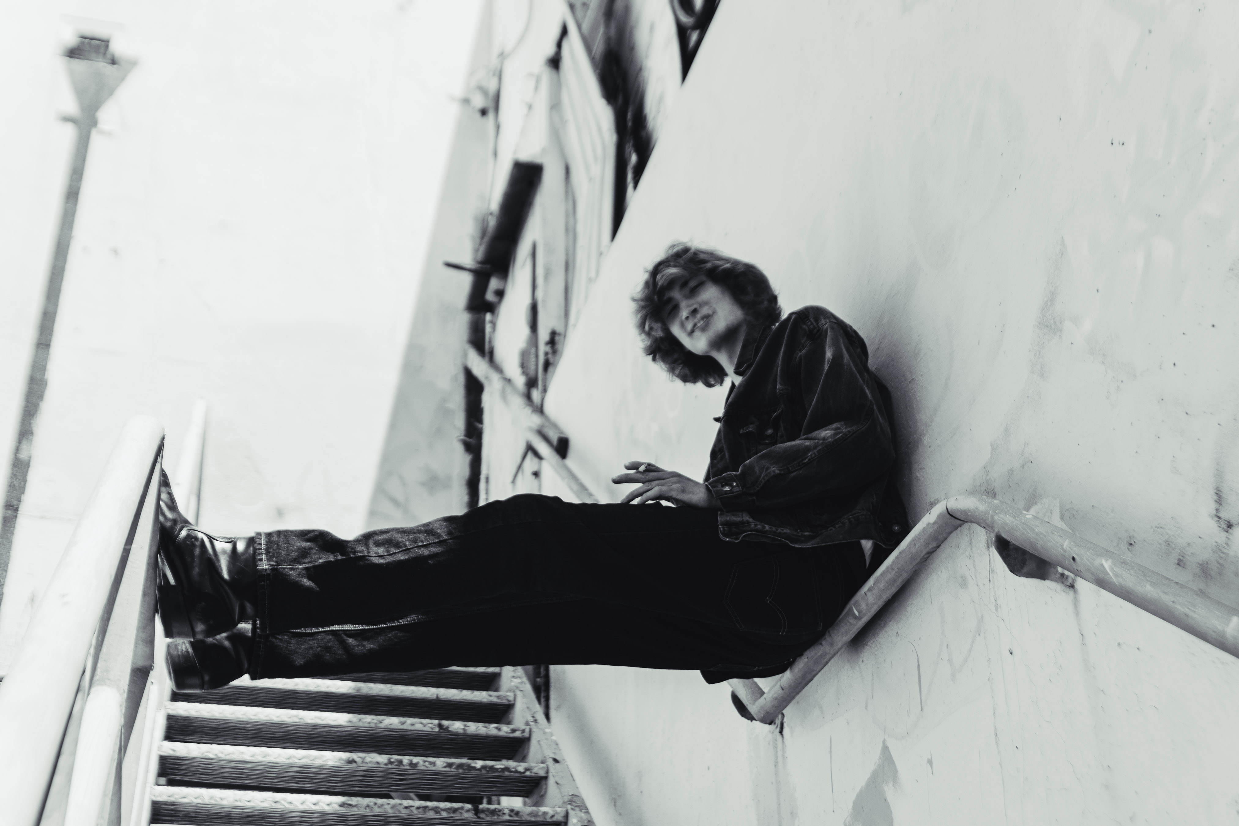 Black and white portrait of an adult sitting on stairs, wearing a jacket and jeans in an outdoor urban environment with walls.