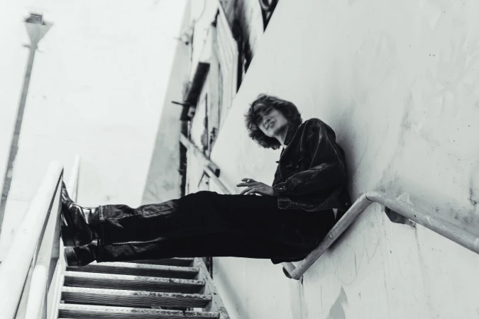 Black and white portrait of an adult sitting on stairs, wearing a jacket and jeans in an outdoor urban environment with walls.