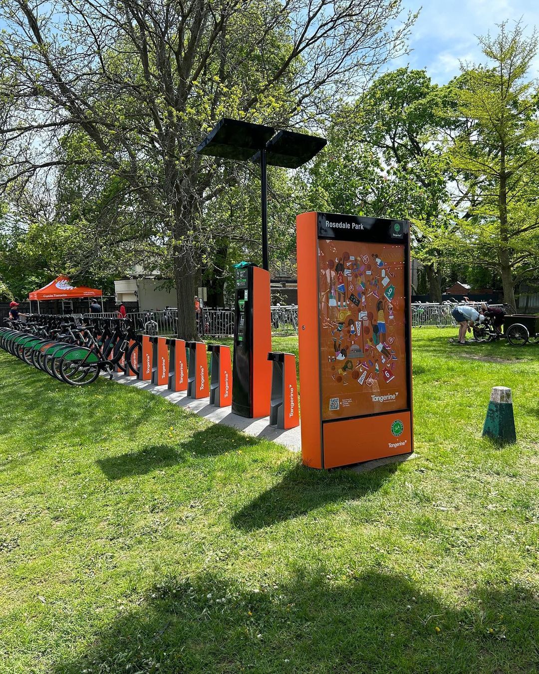 Row of shared rental bicycles parked at a modern docking station with solar panels in a grassy park on a sunny day.