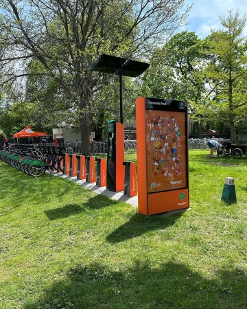 Row of shared rental bicycles parked at a modern docking station with solar panels in a grassy park on a sunny day.