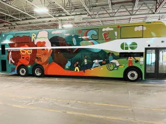 A double decker bus parked inside a large depot, completely covered in a colorful mural with stylized portraits of diverse people.