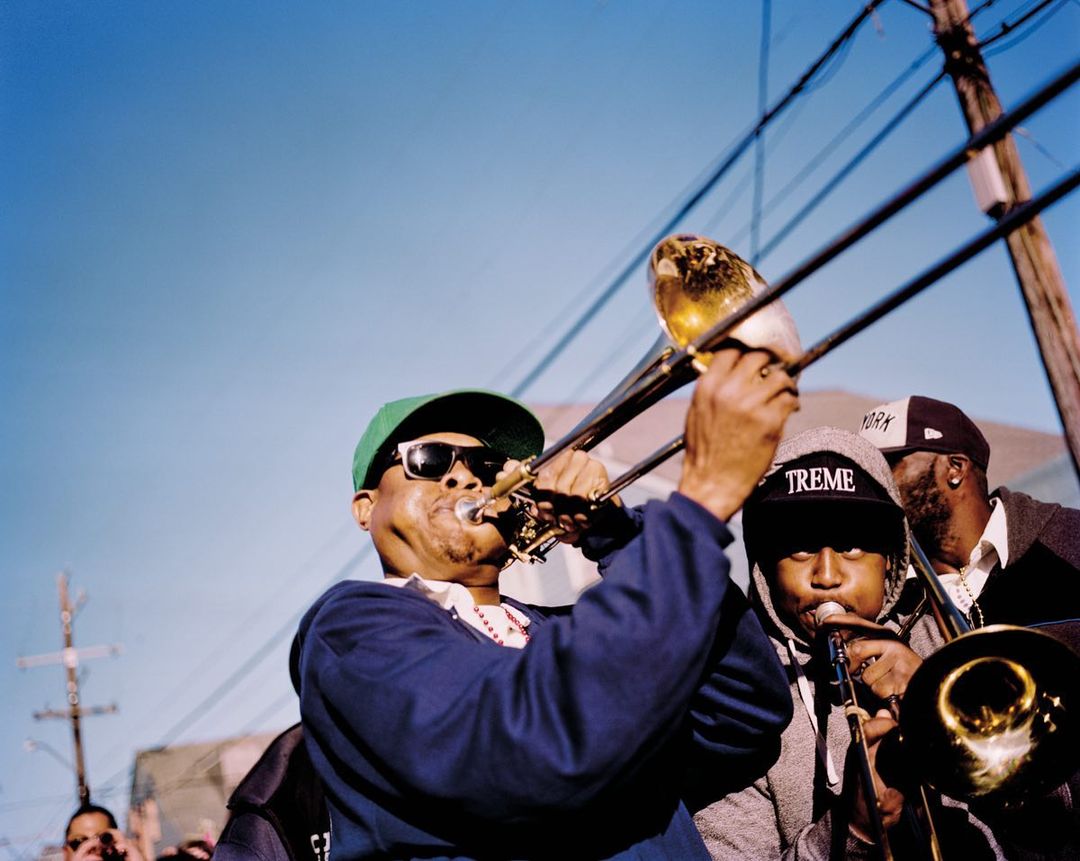 Three musicians playing trombone and trumpet outdoors, wearing hats and sunglasses with a clear blue sky in the background.