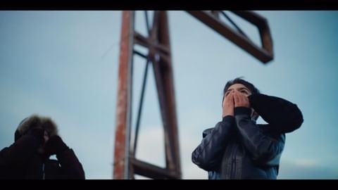 Two people with hands covering their mouths stand outdoors with a metal structure against a blue sky in a cinematic wide shot.