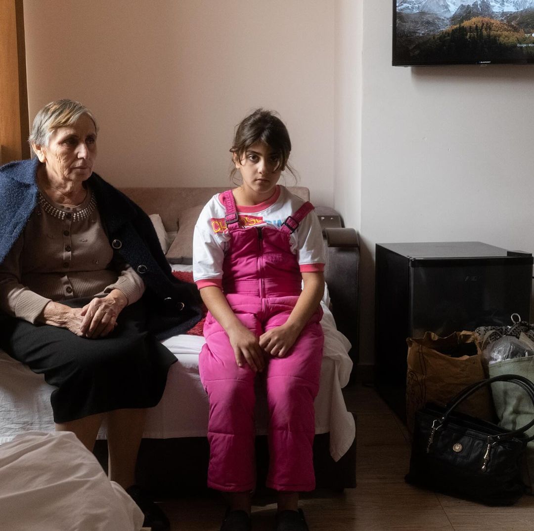 An elderly woman and a young girl sit side-by-side on a bed in a brightly lit room; TV displaying a mountain scene hangs above them.