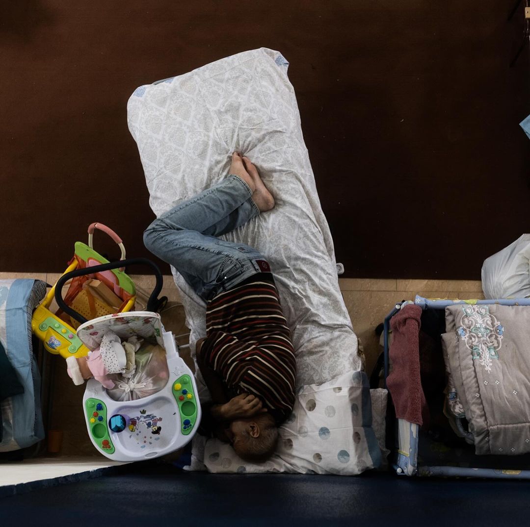 Child in upside down pose on a bed next to baby items. Brown wall and blue floor in the background with overhead camera angle.