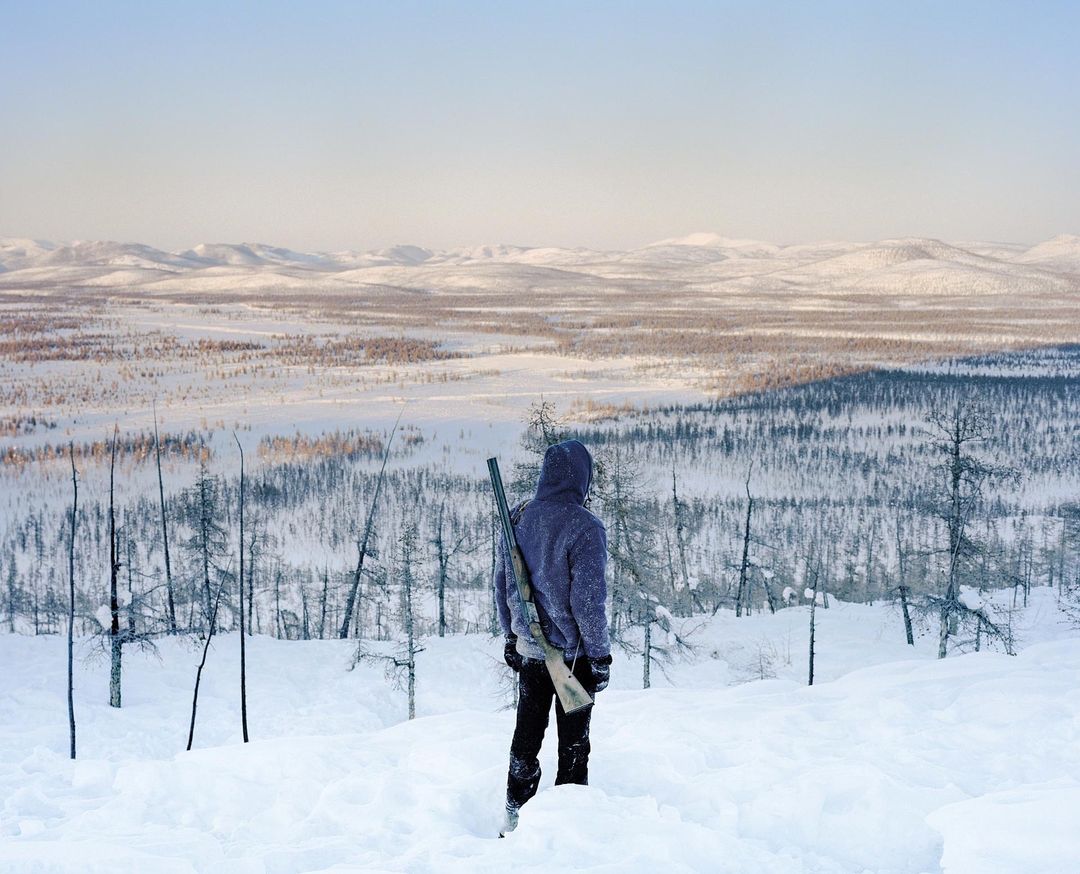 Person with rifle stands in snowy, hilly landscape. Back view of hooded figure in winter forest setting. Cold environment, hunter with gun.