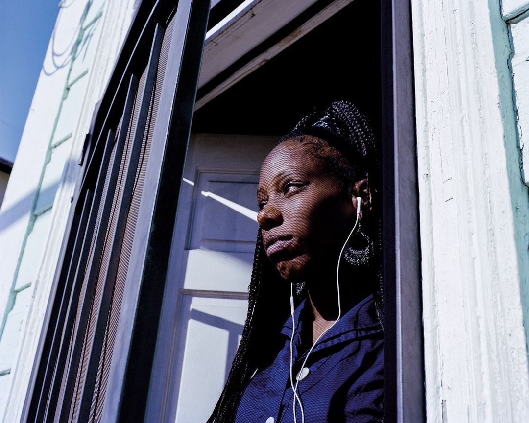 Young Black woman framed by a window with bars, wearing earbuds and a blue shirt, looking to the side in a close-up shot.