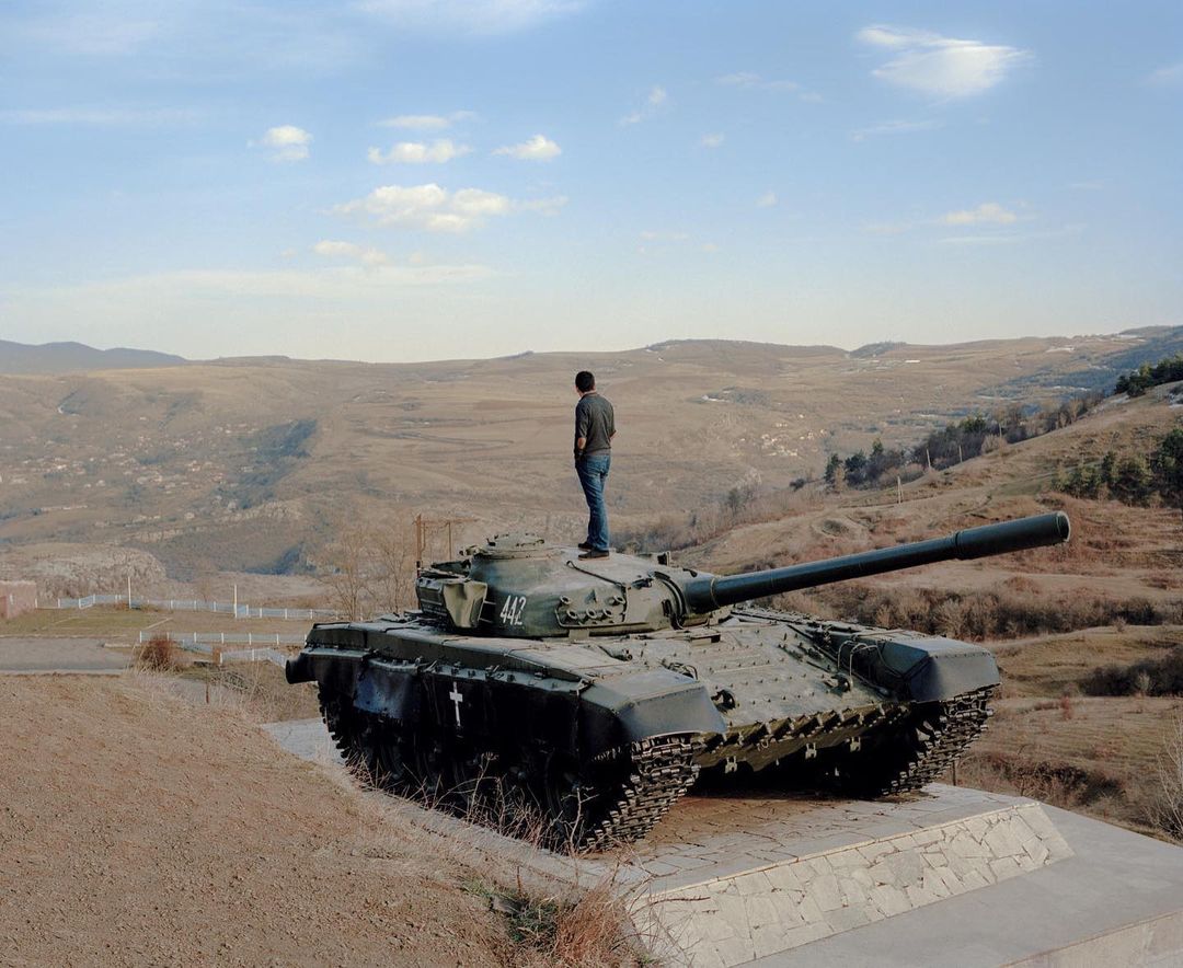 A man stands atop a tank on a stone platform with a mountainous landscape in the background under a clear sky on a sunny day.