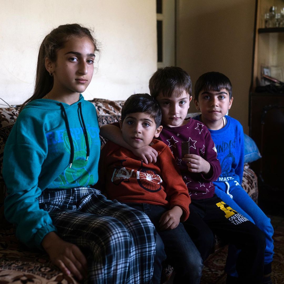 Four young children sit closely together on a patterned sofa in an indoor setting, with neutral wall colors and natural light.