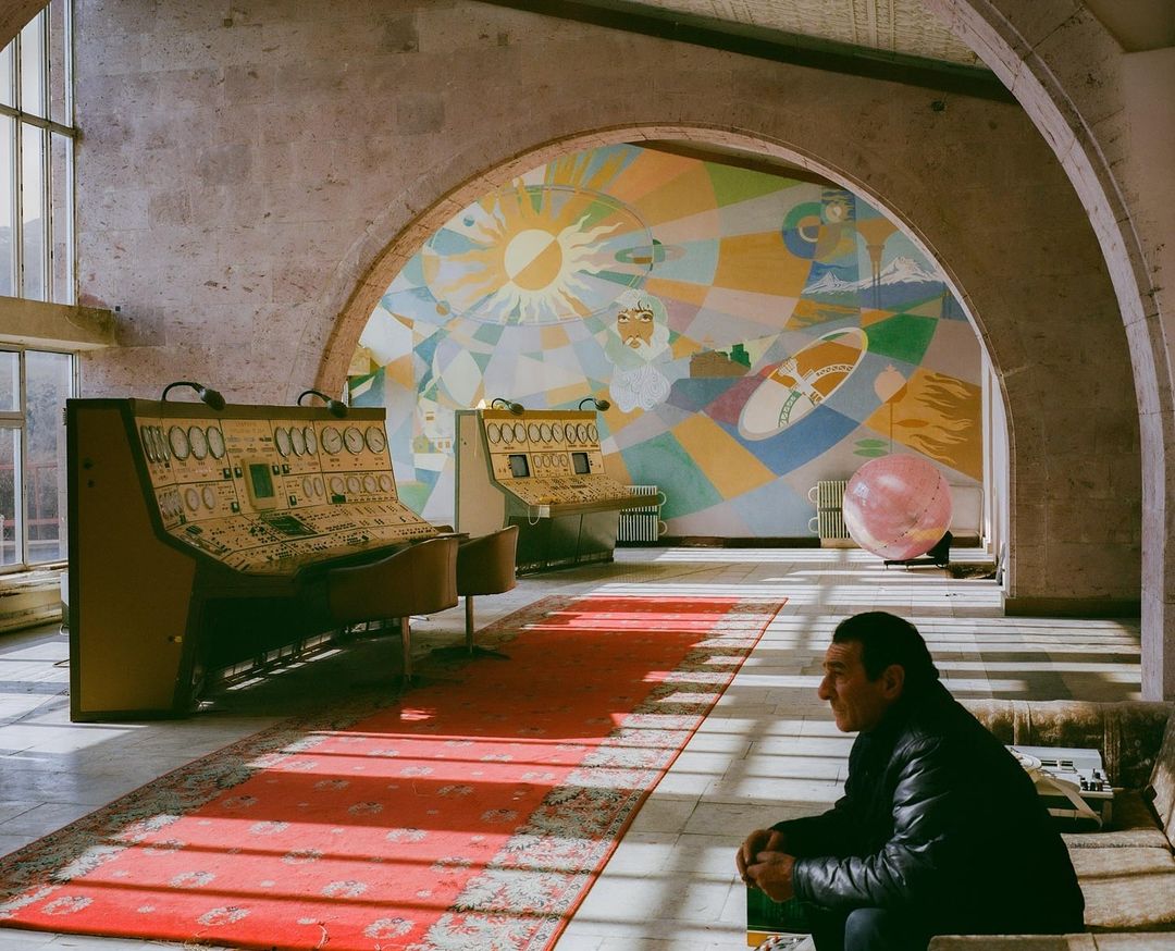 Man sits in grand hall featuring colorful mural, vintage control consoles, red carpet, arches, and large windows casting bright light.