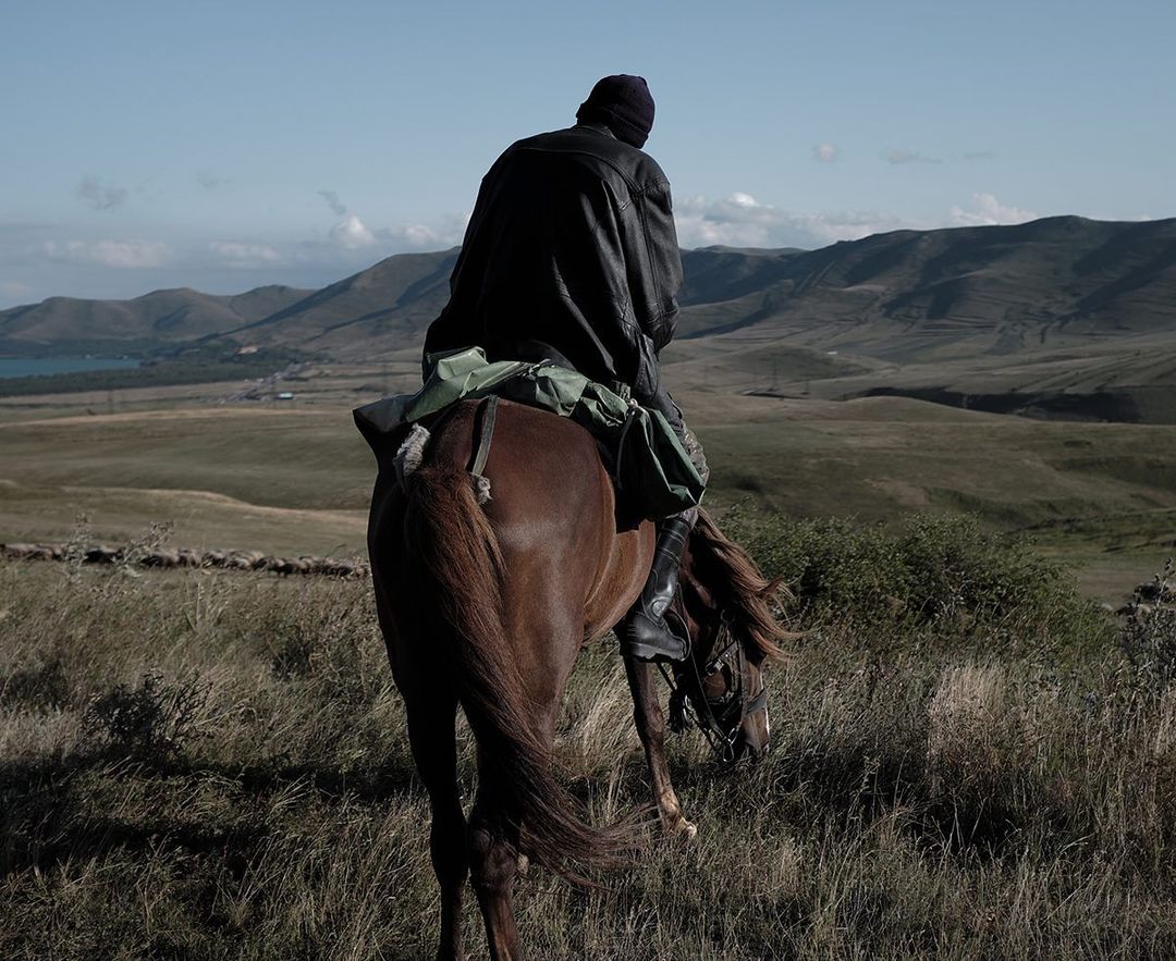 Man on horseback riding through grassy landscape. Hills and lake are visible in background. The person wears dark clothing.