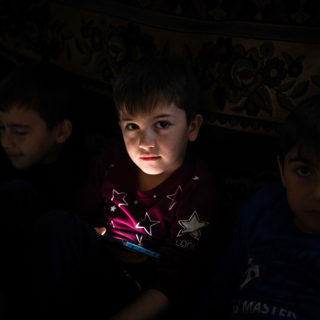 Three boys are captured in a dimly lit, indoor scene, with the focus on one boy holding a device that illuminates his face.