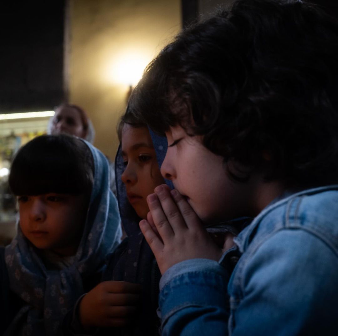 Three young children indoors, one with hands clasped in front of face, another wearing a blue headscarf, somber mood, soft lighting.
