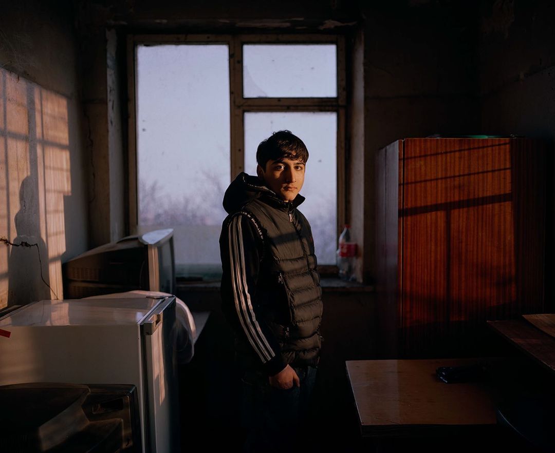 Indoor portrait of a young person in casual wear with an unsmiling expression, standing in front of a window in a dimly lit interior.