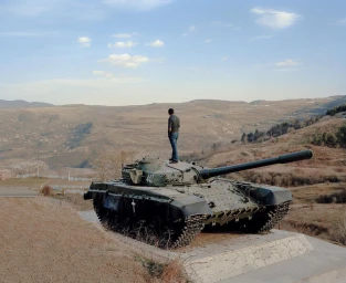 A man standing atop a military tank overlooking a vast hilly landscape under a clear sky, photographed by Alexis Pazoumian.
