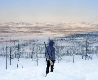 A person in a purple hoodie and dark trousers holding a rifle stands looking over a vast snowy landscape.