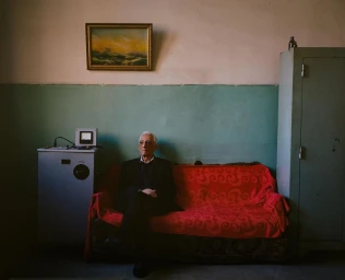 Elderly man seated on a red patterned sofa in a room with a framed landscape painting and metal cabinet